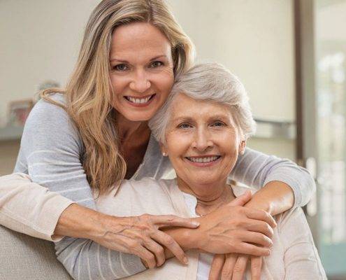 Cheerful mature woman embracing senior mother at home and looking at camera. Portrait of elderly mother and middle aged daughter smiling together. Happy daughter embracing from behind elderly mom sitting on sofa.