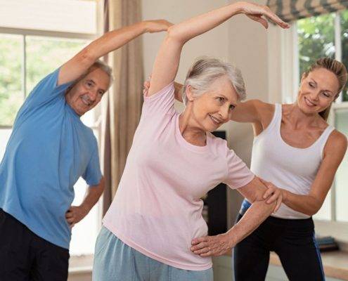 Senior couple doing exercise at home with physiotherapist. Mature gym trainer helping elderly man and old woman exercising at home. Retired husband and wife doing stretching exercises at gym.